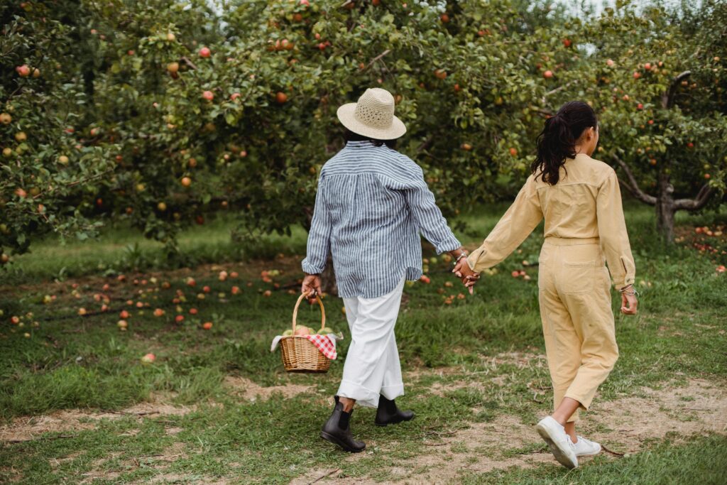 pexels-photo-5528999-5528999 A mother and daughter holding hands while walking through a lush apple orchard with a basket.